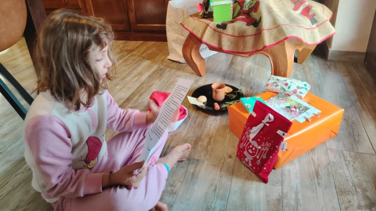 Bambina in pigiama rosa legge una lettera seduta sul pavimento a Natale, accanto a pacchi regalo e all'albero addobbato.