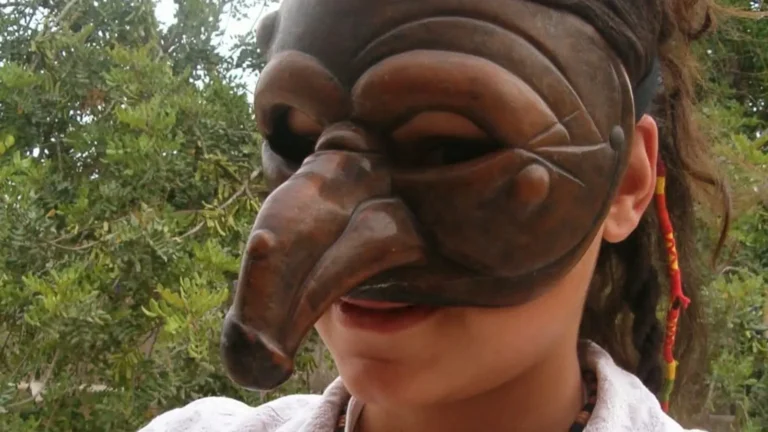 Giovane donna indossa una tradizionale maschera teatrale in legno scolpito con naso prominente, tra alberi all'aperto.