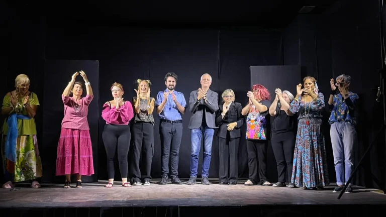 Compagnia di attori sul palcoscenico che applaude e sorride al pubblico durante i saluti finali dello spettacolo teatrale.