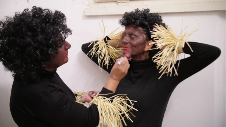 Attrice in costume applica trucco teatrale sul viso di una collega con parrucca riccia dietro le quinte dello spettacolo.