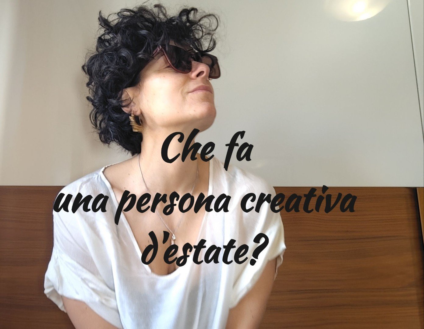 Donna creativa con capelli ricci neri e occhiali da sole, sguardo ispirato verso l'alto, testo overlay: "Che fa una persona creativa d'estate?" – riflessioni su teatro e scrittura estiva