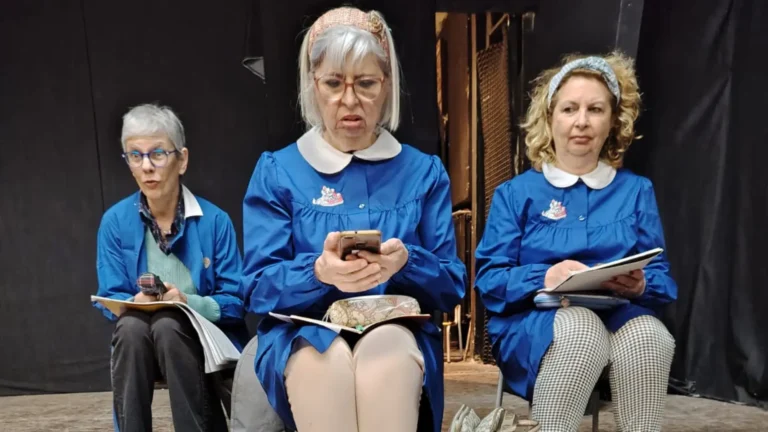 3 attrici col grembiulino blu che interpretano bambine della scuola primaria sedute alle sedioline durante una lezione