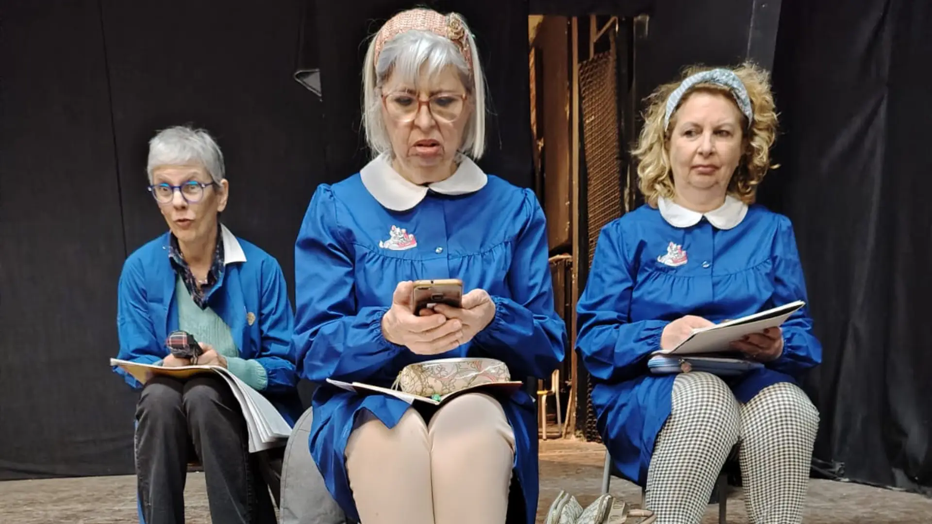 3 attrici col grembiulino blu che interpretano bambine della scuola primaria sedute alle sedioline durante una lezione